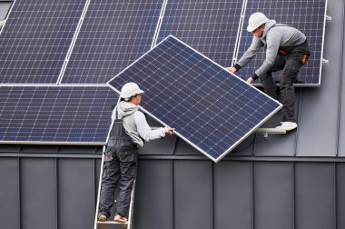 Çatıya güneş paneli kuran çatıcılar. Dışarıda fotovoltaik güneş modülü taşıyan miğferli işçiler. Alternatif ve yenilenebilir enerji kavramı.
