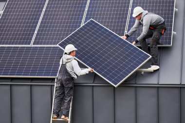 Roofers installing solar panel system on roof of house. Men workers in helmets carrying photovoltaic solar module outdoors. Concept of alternative and renewable energy. clipart