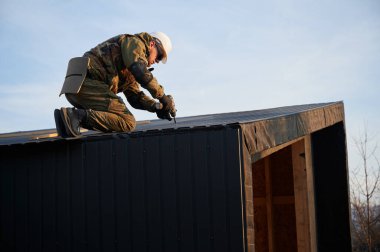 Erkek müteahhit, gelecekteki kulübenin ön cephesi olarak kullanılan siyah kıvrımlı demir çarşaf takıyor. Ahşap çerçeve inşa eden bir işçi. Marangozluk ve inşaat konsepti.