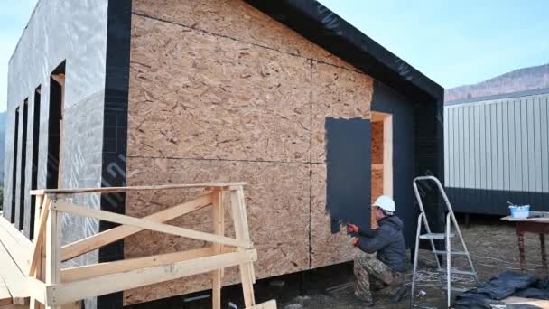 男油漆工用漆辊 用黑色做外漆工作 工人们正在建造木制框架结构房子 木工和建筑概念 — 图库视频影像