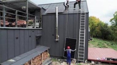 Evin çatısına güneş paneli kuran erkek işçilerin hava görüntüsü. Kasklı elektrikçiler fotovoltaik güneş modülünü açık havada kaldırıyorlar. Alternatif ve yenilenebilir enerji kavramı.