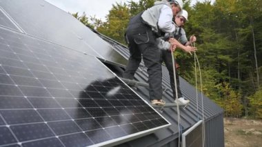 Erkek işçiler evin çatısına güneş paneli kuruyor. Kasklı elektrikçiler fotovoltaik güneş modülünü açık havada halatların yardımıyla kaldırıyorlar. Alternatif ve yenilenebilir enerji kavramı.