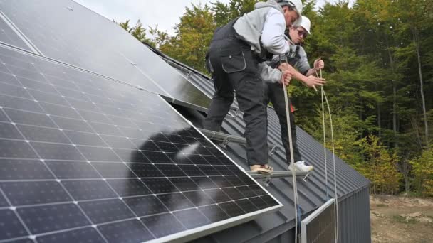 屋根の上にソーラーパネルシステムを設置する男性労働者 屋外ロープの助けを借りて太陽電池モジュールを持ち上げるヘルメットの電気技師 再生可能エネルギーの概念 — ストック動画