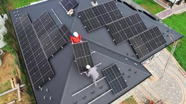 屋根の上に太陽電池パネルシステムを構築する技術者 太陽電池モジュールを屋外に運ぶヘルメットの男性労働者のドローンビュー 再生可能エネルギーの概念 — ストック動画