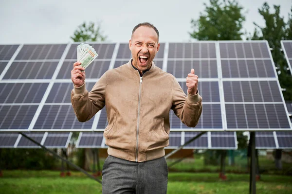 stock image Successful investment in green energy. Man happy about money he earned by investing in alternative energy. Young male shouting cheers with money in hands.