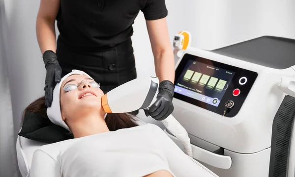 戴安全眼镜的女病人躺在美容设备附近 并进行面部整容 美容院美容师在进行面部治疗时使用激光扫描仪 — 图库照片