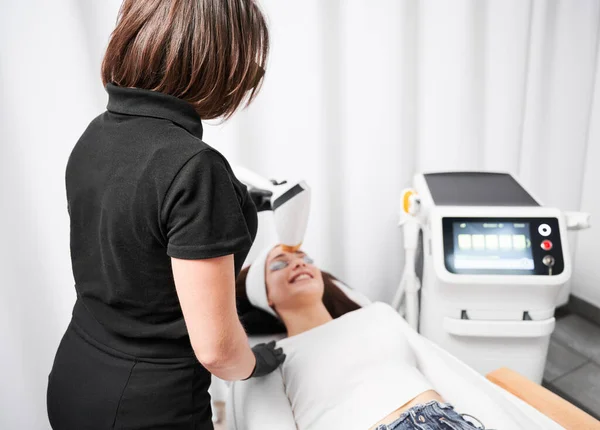 Cosmetologist using diode laser device while performing rejuvenation facial treatment. Woman lying near cosmetology equipment while having face lifting procedure. Focus on female doctor.