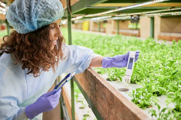 Smiling female gardener in disposable cap holding clipboard and water conductivity meter while standing near shelf with green leafy plants. Woman researcher using digital water tester in greenhouse.