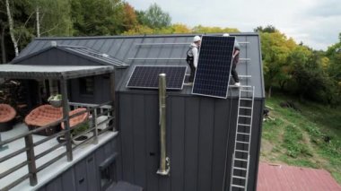 Evin çatısına güneş paneli kuran erkek işçilerin hava görüntüsü. Kasklı elektrikçiler fotovoltaik güneş modülünü açık havada kaldırıyorlar. Alternatif ve yenilenebilir enerji kavramı.