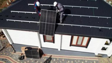 Erkek işçiler evin çatısına güneş paneli kuruyor. Fotovoltaik güneş modülünü kaldıran kasklı elektrikçilerin insansız hava aracı görüntüsü. Yenilenebilir enerji kavramı.