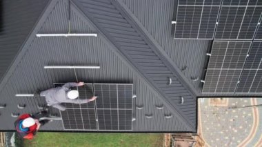 Teknisyenler evin çatısına fotovoltaik güneş panelleri kuruyor. Miğferli erkek mühendislerin güneş modülü sistemi inşa etmelerini gösteren insansız hava aracı görüntüsü. Alternatif, yenilenebilir enerji kavramı.