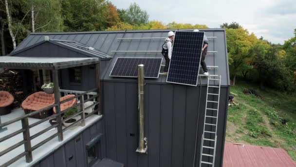 屋根の上に太陽光パネルシステムを設置する男性労働者の空中ビュー ヘルメットの電気技師は屋外で太陽電池モジュールを持ち上げる 再生可能エネルギーの概念 — ストック動画