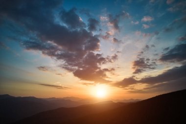 Parlak akşam güneşinin panoramik manzarası ve dağların üzerinde dramatik bulutlu gökyüzü. Renkli akşam gökyüzü altında, bulutlu ve batan güneşli dağ tepelerinin muhteşem manzarası. Doğanın güzelliği konsepti.