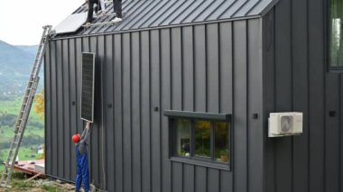 Erkek işçiler evin çatısına güneş paneli kuruyor. Kasklı elektrikçiler fotovoltaik güneş modülünü açık havada halatların yardımıyla kaldırıyorlar. Alternatif ve yenilenebilir enerji kavramı.