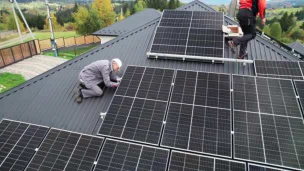 屋根の上に太陽光発電パネルを設置しながらケーブルを接続する電気技師 屋上に太陽電池モジュールを搭載した労働者のドローンビュー 再生可能エネルギーの概念 — ストック動画