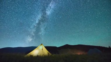 Sarı çadır dağlarda yıldızlı gökyüzüne karşı duruyor. Tepelerde aydınlatılmış çadır. Dağların gece siluetinin arka planında kamp yapmanın parlak ışığı. Zaman aşımı