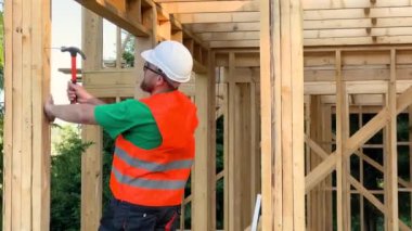 Marangoz ahşap çerçeve inşa ediyor. Gözlüklü sakallı adam binaya çivi çakıyor koruyucu kask ve inşaat yeleği giyiyor. Modern ekolojik yapı kavramı.