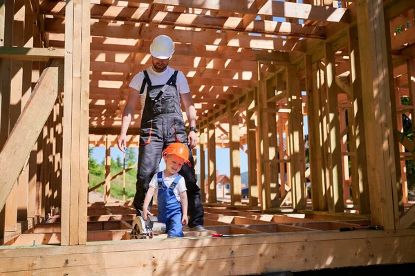 Father with toddler son building wooden frame house. Male worker playing with his son with electric saw on construction site, wearing helmets and overalls on sunny day. Carpentry and family concept.