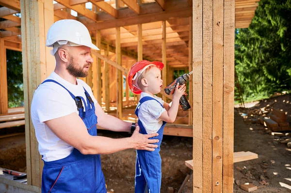 Father with toddler son building wooden frame house. Male builder instructing his son in operation of power drill on construction site, wearing helmets and blue overalls. Carpentry and family concept.