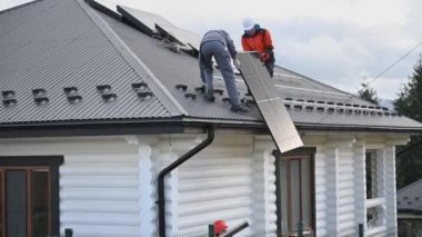 Erkek işçiler evin çatısına güneş paneli kuruyor. Kasklı elektrikçiler fotovoltaik güneş modülünü açık havada kaldırıyorlar. Alternatif ve yenilenebilir enerji kavramı.