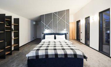 Yatak odasının minimalist iç kısmı büyük yatak odası ve kapalı alanda ahşap kahverengi raflar. Geniş yatak manzaralı, ekose battaniyeli gri duvarlı ve geometrik süslemeli. İç mekan kavramı.