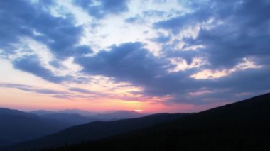 Renkli, dramatik bulutlu gökyüzü altında ağaçlarla çevrili dağ ormanlarının video görüntüleri. Dağlarda, çam ağaçları ve çimenli tepelerle gün batımının resimli manzarası. Doğanın güzelliği konsepti.