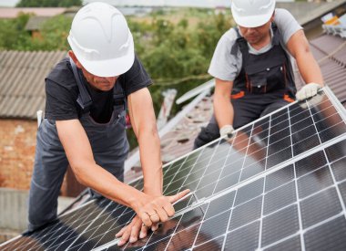 Evin çatısına fotovoltaik güneş modülleri monte eden adamlar. Kasklı elektrikçiler dışarıda güneş paneli sistemi kuruyorlar. Büyü anahtarıyla sıkıyorlar. Alternatif, yenilenebilir enerji kavramı.
