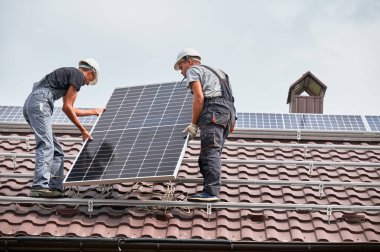Erkek teknisyenler fotovoltaik güneş modülünü evin çatısına kaldırıyorlar. Kasklı elektrikçiler dışarıda güneş paneli sistemi kuruyor. Alternatif ve yenilenebilir enerji kavramı.