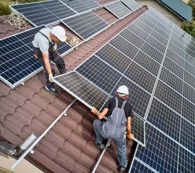 Erkek kurucular evin çatısına fotovoltaik güneş modülleri kuruyorlar. Kasklı mühendisler dışarıda güneş paneli sistemi inşa ediyorlar. Alternatif ve yenilenebilir enerji kavramı. Hava görünümü.