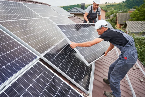 Men workers mounting photovoltaic solar moduls on roof of house. Electricians in helmets installing solar panel system outdoors. Concept of alternative and renewable energy.