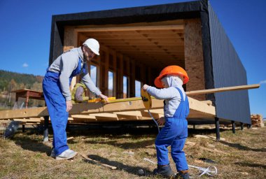 Küçük oğlu olan baba ahşap çerçeve inşa ediyor. İnşaat alanında mezurayla oynayan erkek müteahhit ve çocuk güneşli bir günde kask ve mavi tulum giyiyor. Marangozluk ve aile kavramı.