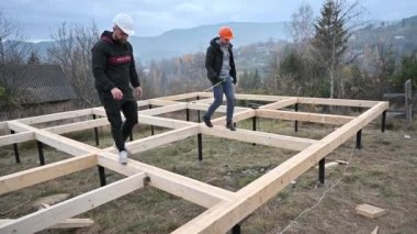 Men workers building wooden frame house on pile foundation. Carpenters using tape measure for measuring wooden planks and making marks with pencil. Carpentry concept.