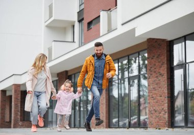 Yeni şehir bölgesinde caddede yürürken küçük bir kızın elini tutan kadın ve erkek. Mutlu ve çocuklu bir aile, apartman yakınlarında geziniyor..