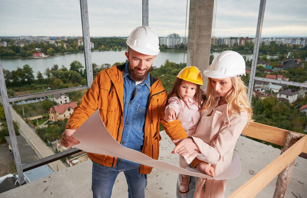 Parents with daughter studying architectural drawings in apartment building under construction. Man holding building plan and smiling while standing next to wife and daughter at construction site.