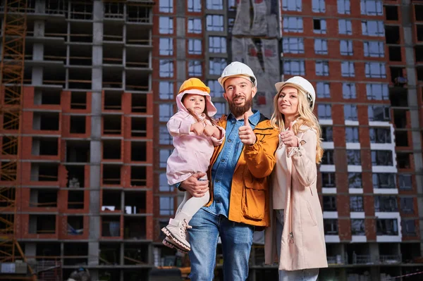 家长和女儿站在室外反对正在建造的公寓楼时 表现出赞许的姿态和微笑 快乐的房主在建筑工地抛下大拇指 — 图库照片