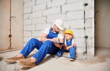 Erkek müteahhit tuğla duvarın yanında oturuyor ve küçük çocuğa belgeleri gösterirken panoya işaret ediyor. Ev tadilatında çalışan, güvenlik kaskı takan ve işçi tulumu giyen bir adam ve çocuk..