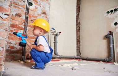 Çocuk inşaat işçisi çömeldi ve kablosuz elektrikli matkapla tuğla duvarı deldi. Bebek kasklı, evde tadilat yaparken matkap aleti kullanıyor..