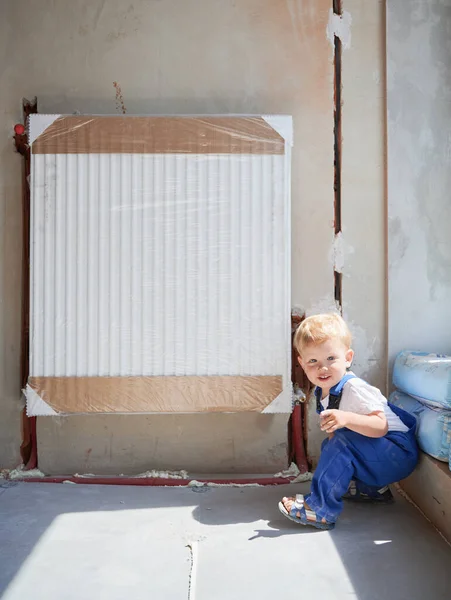 Child plumber in work overalls looking at camera and smiling while crouching down near heating radiator in apartment under renovation. Kid construction worker posing near heating battery at home.