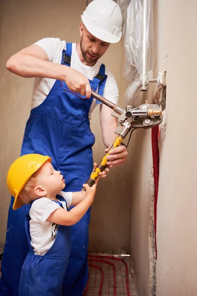 stock image Man and child using pipe pressing tool while installing plumbing pipes in bathroom. Male plumber and kid in safety helmets working on sanitary equipment installation in apartment under renovation.