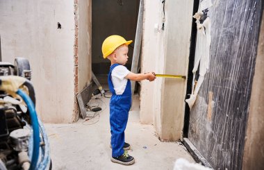 Tadilat altındaki dairede mezura kullanan tam boy bir çocuk inşaat işçisi. Kasklı ve iş tulumlu bir çocuk kapının yanında duruyor ve elinde mezura aleti var..
