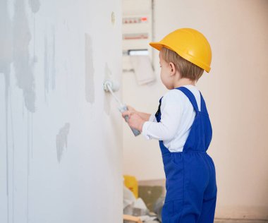 Çocuk inşaat işçisi, tadilat altındaki dairenin duvarlarını boyuyor. Kasklı ve tulumlu bir çocuk evde tadilat yaparken boya silindiri kullanıyor..