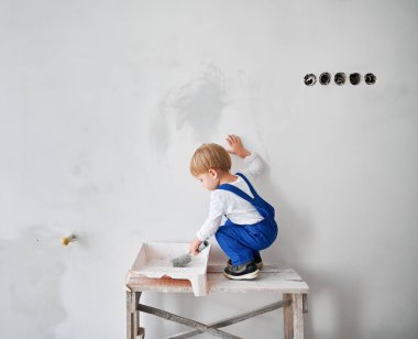 Küçük sevimli inşaat işçisi çocuk daireye beyaz duvar boyuyor. İş yerindeki çocuk ahşap masanın üzerinde duruyor ve evde tadilat sırasında boya silindiri kullanıyor..
