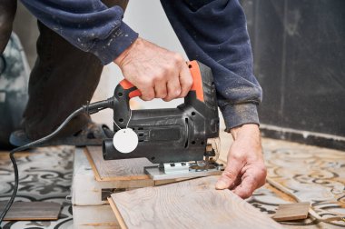 Erkek inşaat işçilerinin elleri yapboz testeresiyle ahşap tahta kesiyor. Elektrik testeresi kullanan bir adam, döşeme kurulumu için kaplama malzemesi hazırlarken....