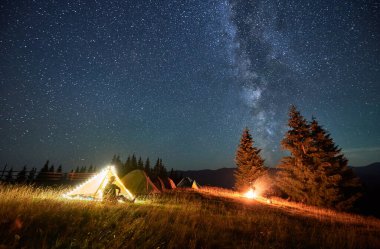 Yıldızlı gökyüzünün altındaki dağlarda gece kampı. Kamp alanındaki çadırın yanında oturan erkek turist, dinleniyor, manzaraya ve ateşe hayran. İnsanoğlu Samanyolu 'yla yıldızlarla dolu güzel gece gökyüzünün tadını çıkarıyor..