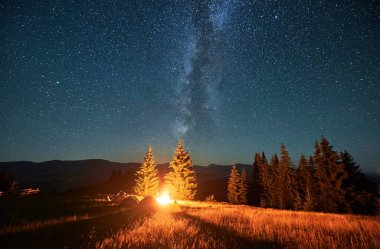 Yıldızlı gökyüzünün altındaki dağlarda gece kampı. Samanyolu ile dolu güzel gökyüzünün altındaki kamp alanında turist çadırları. Yürüyüşçülerin silueti yanan kamp ateşinin yanında dinleniyor. Turizm kavramı.