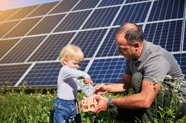 小儿子和爸爸一起存钱 把钱存进储蓄罐 爸爸教小孩如何为将来存钱 把他们的积蓄放在太阳能电池板的基础上 — 图库照片