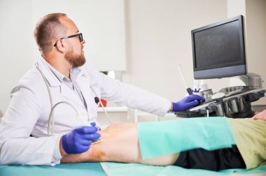 Doktor ultrason testi yapıyor, hasta kanepede yatıyor. Üniformalı ve gözlüklü bir erkek uzman, ekrana bakıyor. Tıp ve sağlık hizmetleri kavramı.