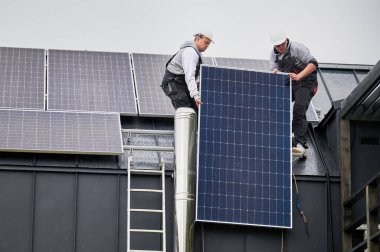 Erkek işçiler evin çatısına güneş paneli kuruyor. Kasklı elektrikçiler fotovoltaik güneş modülünü açık havada halatların yardımıyla kaldırıyorlar. Alternatif ve yenilenebilir enerji kavramı.