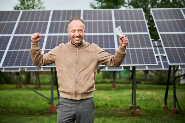 Man happy about money he earned by investing in alternative energy. Successful investment in green energy. Young male shouting cheers with money in hands.