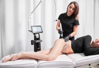 Doktor kozmetik uzmanı, kadın kalçasında deri sıkılaştırma tedavisi uygularken mikronikleme cihazı kullanıyor. Gerilme izleri çıkarma işlemi sırasında tıbbi sandalyeye uzanmış gülümseyen kadın..